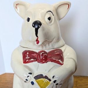 May include: A vintage ceramic cookie jar shaped like a white bear with a red bow tie. The bear holds a yellow and brown object. The jar has black eyes, nose, and paw details. A charming kitchen collectible.