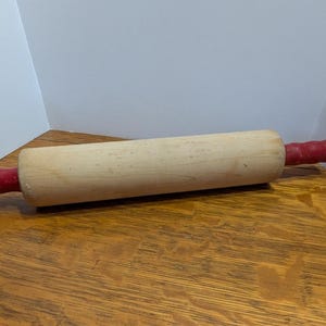 May include: A wooden rolling pin with red handles. The rolling pin is made of light wood and has a smooth surface.