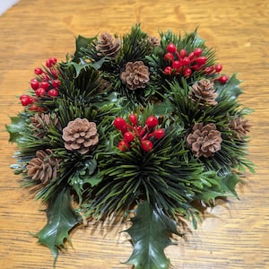 May include: A circular Christmas decoration featuring green pine needles, brown pine cones, red berries, and green holly leaves. The arrangement is set against a wooden surface, creating a festive holiday display.