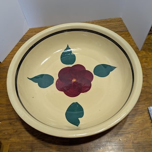 May include: A cream-colored ceramic bowl with a dark rim and a floral design. The flower is a deep red with a yellow center, surrounded by four green leaf shapes. The bowl is likely used for serving.