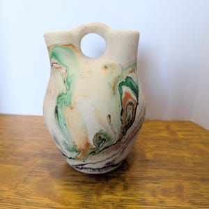 May include: A ceramic vase with a rounded body and a central circular handle at the top. The surface features a marbled pattern with swirls of cream, sage green, burnt orange, and black colors against a matte, off-white textured base.