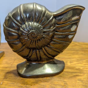 May include: A bronze-colored, nautilus shell sculpture. The shell is detailed with spiral ridges and sits on a rectangular base. The sculpture is displayed on a wooden surface.