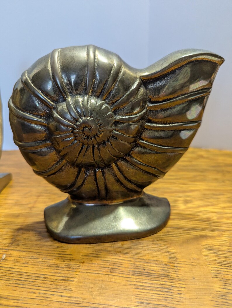 May include: A bronze-colored, nautilus shell sculpture. The shell is detailed with spiral ridges and sits on a rectangular base. The sculpture is displayed on a wooden surface.