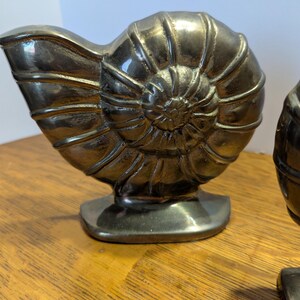 May include: Two metallic silver-colored nautilus shell sculptures. The sculptures are detailed with spiral patterns and sit on a rectangular base. The shells are displayed on a wooden surface.