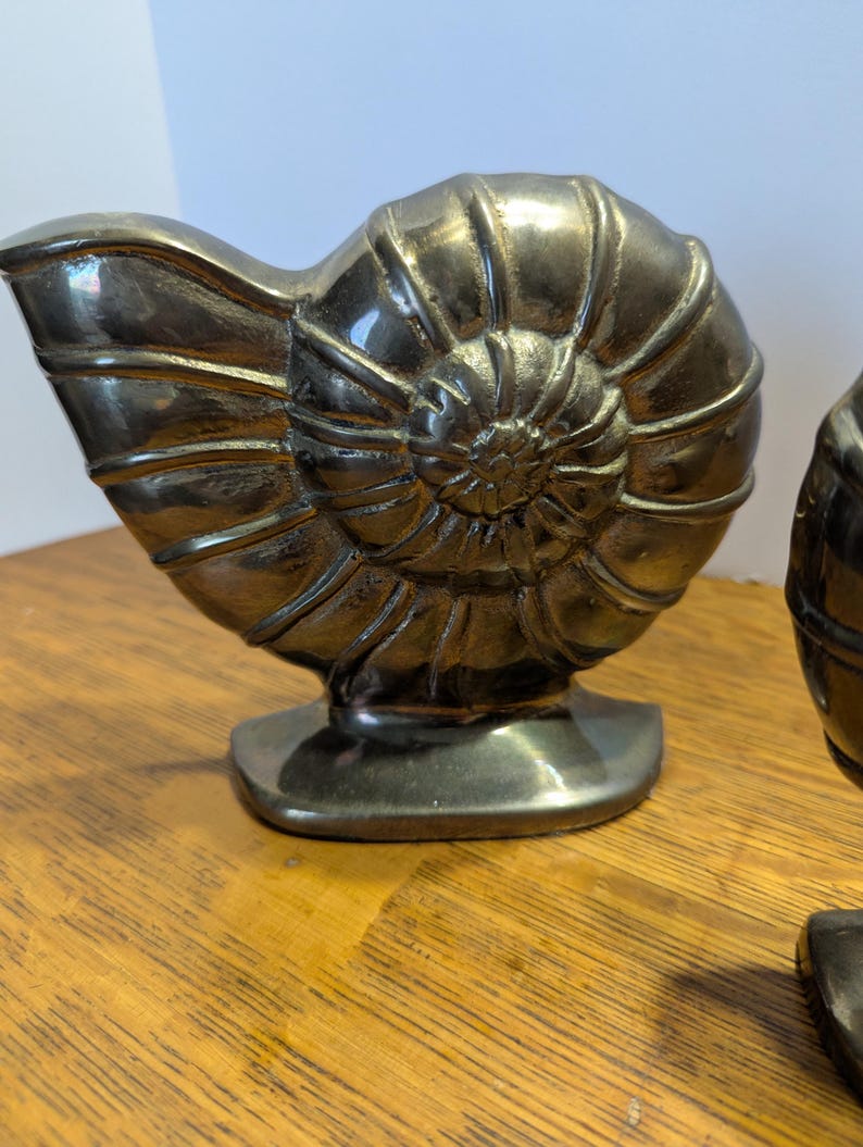 May include: Two metallic silver-colored nautilus shell sculptures. The sculptures are detailed with spiral patterns and sit on a rectangular base. The shells are displayed on a wooden surface.