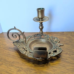May include: An antique brass candle holder with an ornate, oval base and a single candle cup. The base features intricate scrollwork and a small, rectangular match holder. A decorative swirl extends from the side.