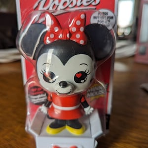 May include: A red and white Popsies Minnie Mouse character toy. The toy is a bobblehead with a red dress and black shoes. The toy is packaged in a clear plastic box with a red and white background. The box has the words "Popsies" and "Minnie Mouse" on it.