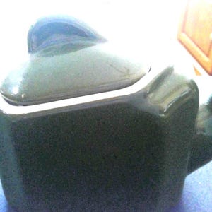 May include: A dark green ceramic teapot with a matching lid. The teapot has a square shape with rounded edges, a curved handle, and a spout. The lid has a small knob on top. The teapot is sitting on a blue surface.