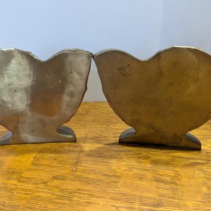 May include: Two antique brass heart-shaped bookends. The bookends have a textured surface and a warm, golden hue. They are placed side-by-side on a wooden surface, showcasing their unique design and vintage appeal.