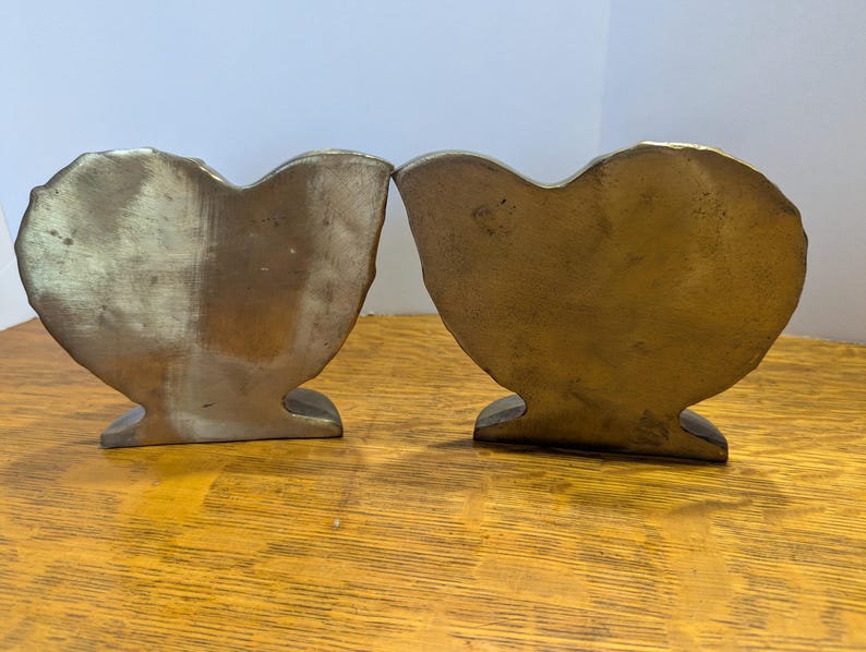 May include: Two antique brass heart-shaped bookends. The bookends have a textured surface and a warm, golden hue. They are placed side-by-side on a wooden surface, showcasing their unique design and vintage appeal.