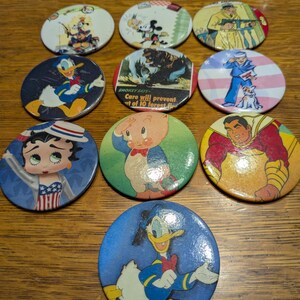 May include: Nine round, colorful, vintage cartoon character buttons. The buttons feature characters like Mickey Mouse, Donald Duck, Porky Pig, and Betty Boop.  The buttons are in various colors, including red, blue, yellow, and green.