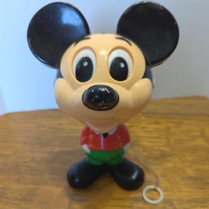 May include: Vintage Mickey Mouse toy figurine. The toy features a tan face, black ears, and a red shirt with green pants. The toy has a string attached to a white ring.