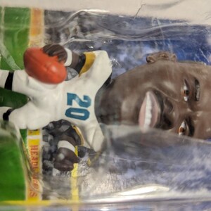 May include: A plastic figurine of a football player wearing a white and teal jersey with the number 20 on it. The player is holding a football and is standing on a green base with the word "SAINTS" printed on it.