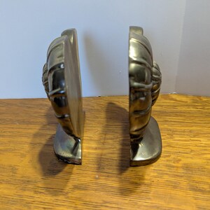 May include: Two brass-colored bookends, each featuring a detailed, textured design. The bookends are positioned side-by-side, showcasing their decorative front. They are placed on a wooden surface, with a neutral background.