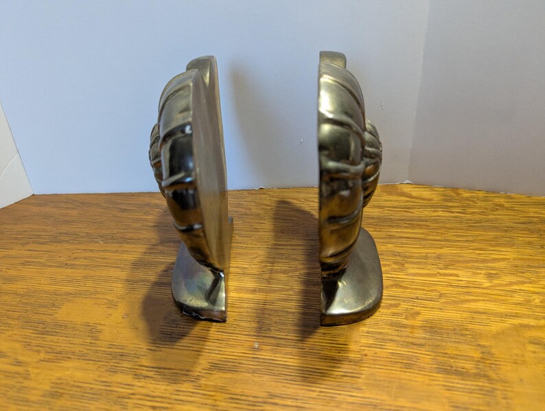 May include: Two brass-colored bookends, each featuring a detailed, textured design. The bookends are positioned side-by-side, showcasing their decorative front. They are placed on a wooden surface, with a neutral background.