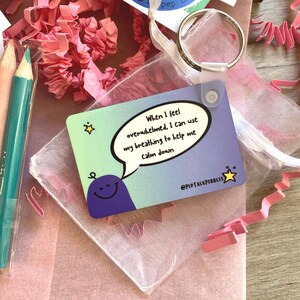 Puede incluir: Una tarjeta azul y morada con un personaje de dibujos animados y un globo de di&aacute;logo que dice "Cuando me siento abrumado, puedo usar mi respiraci&oacute;n para ayudarme a calmarme." La tarjeta est&aacute; en una peque&ntilde;a bolsa de malla blanca con un anillo plateado.