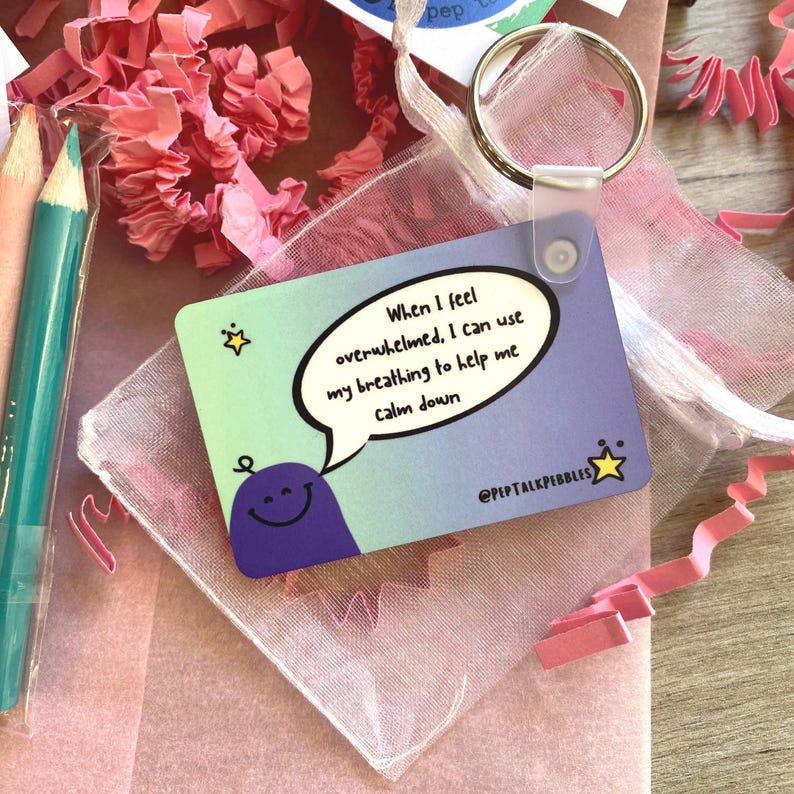 Puede incluir: Una tarjeta azul y morada con un personaje de dibujos animados y un globo de di&aacute;logo que dice "Cuando me siento abrumado, puedo usar mi respiraci&oacute;n para ayudarme a calmarme." La tarjeta est&aacute; en una peque&ntilde;a bolsa de malla blanca con un anillo plateado.