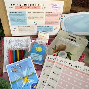 May include: A pink and white box filled with self-care items for positive mental health. The box includes coloring cards and pencils, a worry duck, a coconut foot mask, and cards with positive affirmations and quotes. The box also includes a 5-day planner with prompts for well-being.
