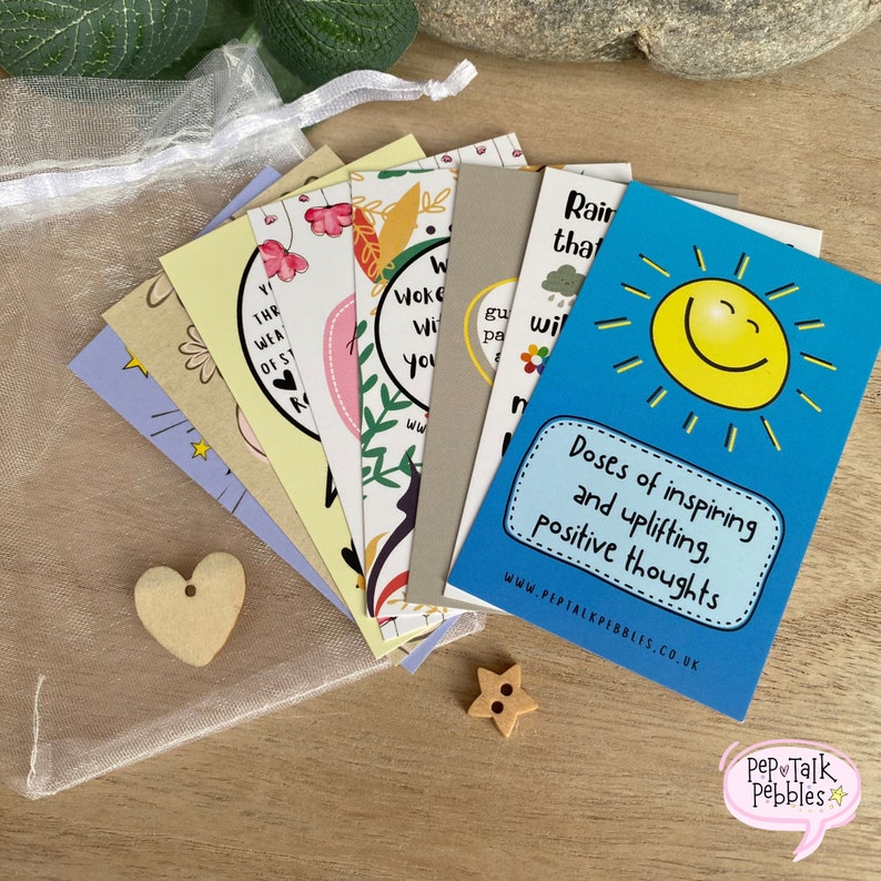 May include: A set of colorful affirmation cards with inspirational messages and a cheerful sun design. The cards are in a white mesh bag with a wooden heart and star. The text on the cards includes "Doses of inspiring and uplifting, positive thoughts" and "www.peptalkpebbles.co.uk".