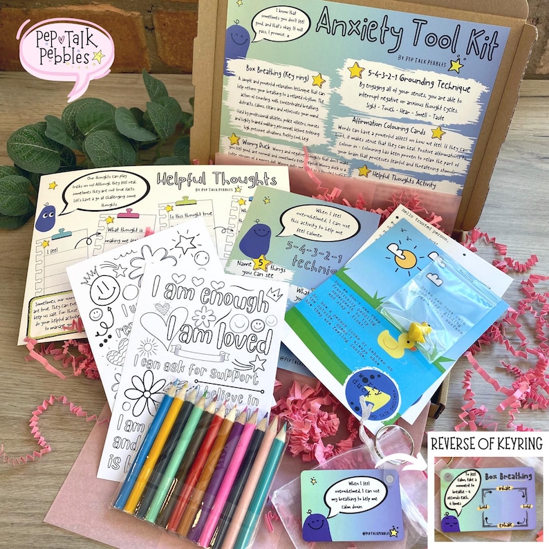 Puede incluir: Una caja de regalo rosa y blanca llena de herramientas para aliviar la ansiedad. La caja incluye p&aacute;ginas para colorear con afirmaciones positivas, un llavero con un ejercicio de respiraci&oacute;n y un juego de l&aacute;pices de colores. La caja est&aacute; etiquetada como "Anxiety Tool Kit" y presenta un personaje de dibujos animados.