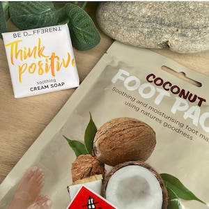 May include: A white bar of soap with the text "BE D_FFERENT Think Positive soothing CREAM SOAP" on it. A brown paper packet with the text "COCONUT FOOT PAC Soothing and moisturising foot mask using natures goodness" on it. The packet has a picture of a coconut and a green leaf.
