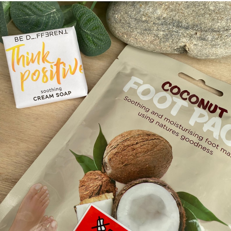 May include: A white bar of soap with the text "BE D_FFERENT Think Positive soothing CREAM SOAP" on it. A brown paper packet with the text "COCONUT FOOT PAC Soothing and moisturising foot mask using natures goodness" on it. The packet has a picture of a coconut and a green leaf.