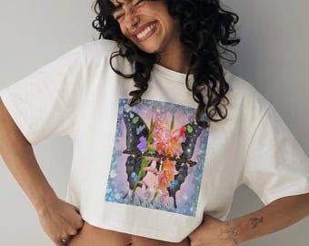 May include: White crop top featuring a butterfly design with floral accents and the text "dancing the fire of suffering". The butterfly has black wings with colorful details. The shirt is a casual style.