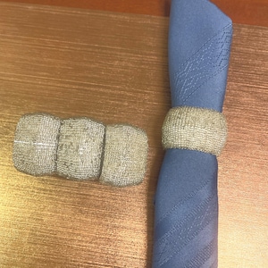 May include: A blue folded napkin with a beaded napkin ring and a set of three beaded napkin holders. The napkin ring and holders are silver in colour. The items are on a gold-coloured wooden surface.