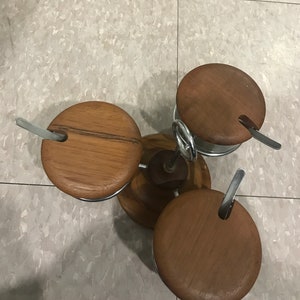 Puede incluir: Un juego de tres recipientes para condimentos de madera con tapas y cucharas. Los recipientes están hechos de madera con un acabado natural y tienen una base de metal.