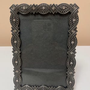 May include: A rectangular silver-toned picture frame with an ornate, repeating pattern. The frame is embellished with small, clear crystals. The interior of the frame is a dark gray color, and the frame is standing upright.