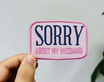 Puede incluir: Un parche bordado rectangular con esquinas redondeadas, fondo rosa claro y borde rosa oscuro. El texto SORRY está bordado en letras grandes de color azul oscuro, con las palabras ABOUT MY HUSBAND en letras rosas más pequeñas.
