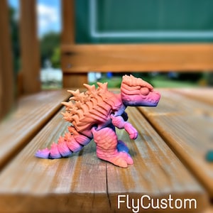 May include: A small, pink and orange 3D printed dinosaur toy. The dinosaur is posed in a walking position with its head turned to the side. The toy is on a wooden surface.