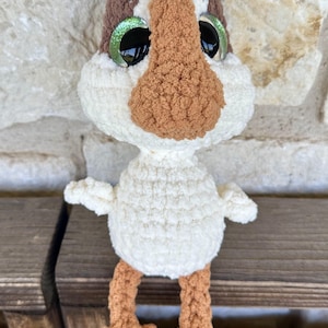 Puede incluir: Una muñeca de pájaro de ganchillo hecha a mano con cuerpo blanco, pico marrón y cabeza marrón y blanca. La muñeca tiene grandes ojos verdes brillantes y pies marrones. Está sentada sobre una superficie de madera.