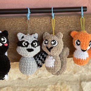 May include: Four crocheted animal keychains hanging from a black rod. The animals are a skunk, a raccoon, a squirrel, and a fox. The skunk is black and white, the raccoon is gray and black, the squirrel is brown and white, and the fox is orange and white.