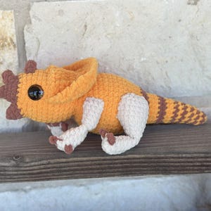 Puede incluir: Un dinosaurio Triceratops de ganchillo hecho a mano en tonos naranja, marrón y crema. El peluche presenta una cabeza detallada con cuernos, ojos negros grandes y una cola a rayas. Está apoyado sobre una superficie de madera.