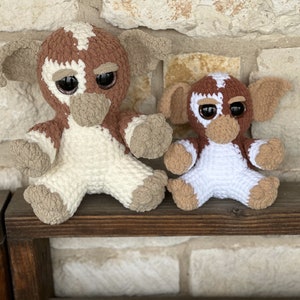 Puede incluir: Dos juguetes de peluche tejidos a crochet, uno marrón y blanco y otro más pequeño blanco y marrón, con ojos grandes y una nariz cosida. Los juguetes están sentados en un estante de madera.