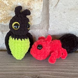 May include: Two crocheted stuffed animals, one black with green accents and one red. Both have large black eyes and are sitting on a wooden surface.
