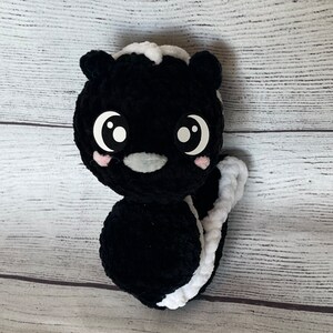 May include: A black and white crocheted skunk plush toy with large eyes and a pink blush. The toy has a long, white tail.
