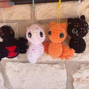 May include: Four crocheted plush toys hanging from a string. The toys are a black spider with red accents, a white ghost with colourful sprinkles, an orange bat with large eyes, and a black cat with colourful sprinkles.