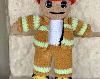 May include: A crocheted plush toy firefighter doll wearing a red hat with a black brim and a white number "1" on the front. The doll has brown skin, blue eyes, and a brown crocheted uniform with green stripes.