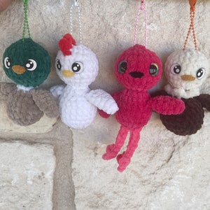 Op de afbeelding: Vier gehaakte pluche speelgoedjes die aan touwtjes hangen. De speelgoedjes zijn een groene eend, een witte kip, een roze flamingo en een bruin-witte vogel.