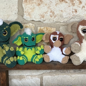 May include: Four crocheted plush toys, inspired by the movie Gremlins, are sitting on a wooden shelf. The toys are green, brown, and white. The toys have large eyes and are in a sitting position.