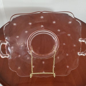 May include: A clear pink glass serving plate with a scalloped edge and two handles. The plate features etched star designs and is displayed on a gold-colored metal stand. The plate is approximately 10 inches in diameter.