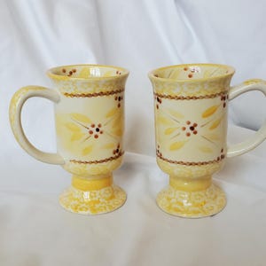 May include: Two ceramic mugs with a cream-colored base and yellow floral design. The mugs have a pedestal base, a curved handle, and a decorative border. The floral design includes yellow leaves and brown dots. The mugs are suitable for hot beverages.