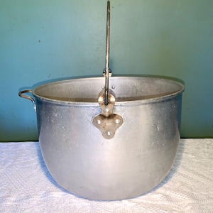 May include: A well-used, vintage aluminium cooking pot with a curved body and a long, arched handle. It has a small handle on the side. The pot is set against a blue-green background, suggesting it is an older item.