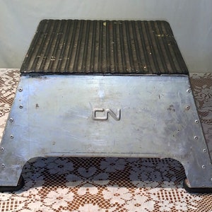 Puede incluir: Un taburete de metal plateado con una parte superior negra acanalada. El taburete tiene un logotipo CN en la parte delantera.