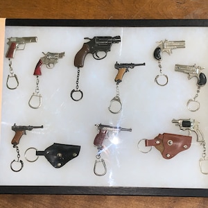 Op de afbeelding: Een vitrine met een verzameling van 10 miniatuur pistool sleutelhangers. De sleutelhangers zijn gemaakt van metaal en hebben verschillende stijlen, waaronder revolvers, pistolen en geweren. Sommige sleutelhangers hebben bruine houten handvatten.