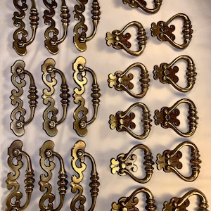May include: Set of 20 antique brass cabinet drawer pulls with ornate designs. The pulls have a decorative, scrolled design and a heart-shaped opening.