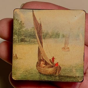 May include: A small, square, vintage wooden box featuring a painted nautical scene. The artwork shows sailboats on water with figures in the boats and on the shore. The colour palette includes muted greens, browns, and blues.