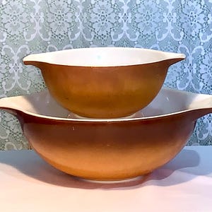 Puede incluir: Dos cuencos de mezcla Pyrex vintage. Los cuencos son marrones con bordes y asas blancos. El cuenco más grande está en la parte inferior y el cuenco más pequeño está apilado encima. Los cuencos son de vidrio y tienen un diseño clásico.